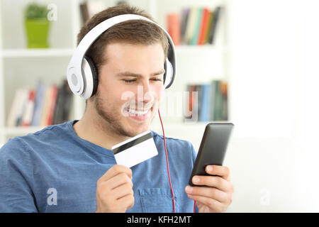 Happy guy wearing headphones paying streaming music with credit card and a smart phone sitting on a sofa at home Stock Photo