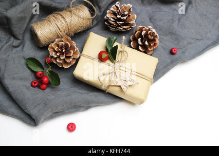 Christmas festive styled stock image composition. Handmade Christmas gift box, red berries, pine cones isolated on grey linen blanket. Flat lay, top view. Winter holiday background. Winter concept. Stock Photo