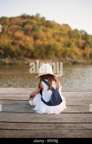 Little girl sitting alone on a wooden bridge by the river Stock Photo
