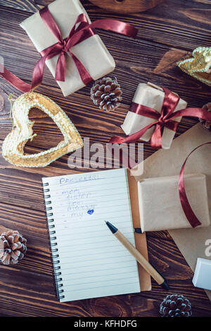 Top view Holiday decorations and notebook with list of gifts on wooden rustic table, flat lay style. Christmas planning concept. Selective focus. Spac Stock Photo