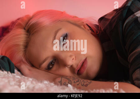 Portrait of young woman with pink hair, lying down, closeup, looking directly at the camera, head resting on tattooed forearm. Stock Photo