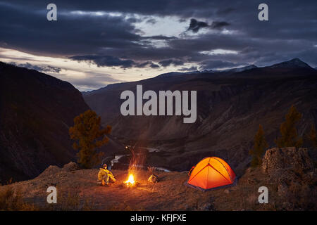 A man near illuminated tent and campfire in mountains in dawn Stock Photo