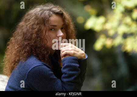 Candid natural portrait of a young woman with long curly brown hair and tranquil thoughtul expression on her face in fall nature scenery Stock Photo
