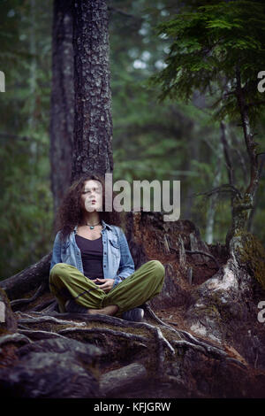 Young woman meditating in a forest sitting in meditation pose with her legs crossed leaning with her back against a tree in beautiful tranquil calm na