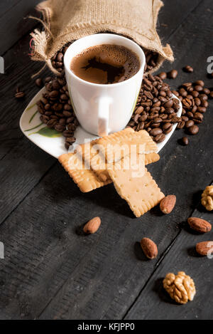 Cup of coffee and coffee beans on a plate with roasted almonds, walnuts and biscuits on a black wooden table Stock Photo