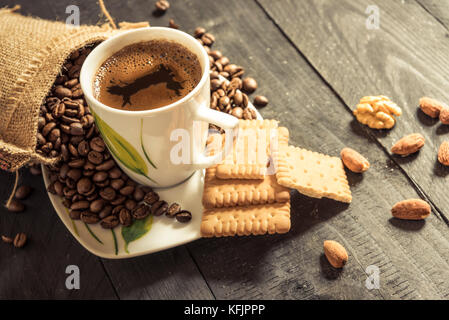 Coffee cup on a plate and coffee beans pouring out from a burlap sack, biscuits, walnuts and almonds on a wooden table Stock Photo