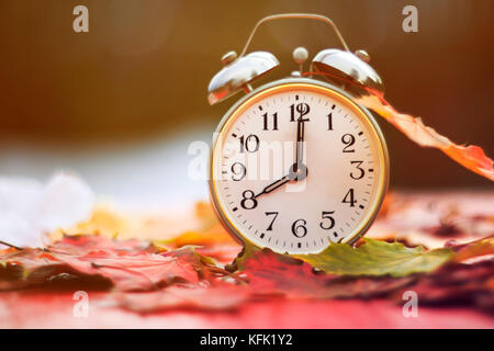 old metal clock standing on a red wooden table amongst fallen autumn leaves a bright Sunny autumn morning Stock Photo