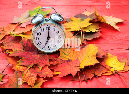 old metal clock standing on a red wooden table amongst fallen autumn leaves a bright Sunny autumn morning Stock Photo