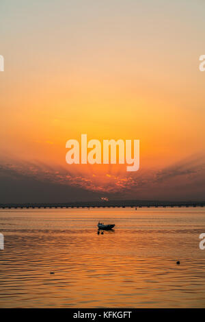 A fishing boat at sunset, with a bridge on the horizon. Fiery sky, with rays of light coming behind clouds Stock Photo