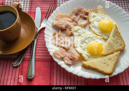 Delicious Breakfast - a Cup of coffee, a plate of fried eggs, bacon and toast, next to the Cutlery on red checkered napkin. Stock Photo