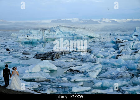 Bride in wedding dress and groom holding hands looking at blue iceberg lake at Jokursarlon glacial lagoon glacier tourist attraction Southern Iceland Stock Photo