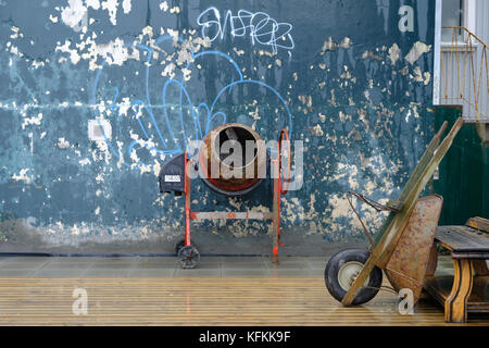 Old red cement concrete mixer and brown rusted wheelbarrow leaning against wooden bench with peeling paint blue wall and graffiti in background Stock Photo