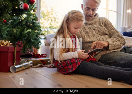 Little girl with grandfather sitting by christmas tree at home and using digital tablet. Both sitting on floor in living room and looking at tablet pc Stock Photo