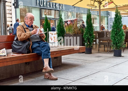 Elderly, senior Caucasian man wearing glasses, old age pensioner seated with crossed legs on a bench, reading papers, downtown Vienna, Austria, Europe Stock Photo