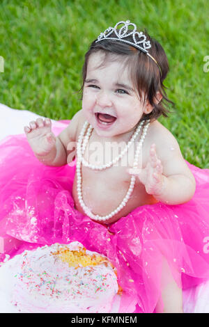 Smiling happy baby toddler girl first birthday anniversary party. Ecstatic and laughing, raised hands, face dirty from pink cake. Princess costume Stock Photo