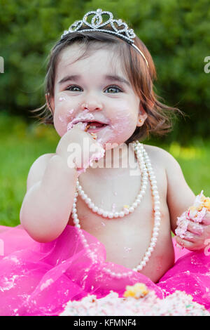 Smiling happy baby toddler girl first birthday anniversary party. Licking fingers with face dirty from pink cake. Princess tiara costume Stock Photo