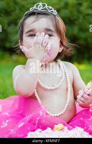 Smiling happy baby toddler girl first birthday anniversary party. Licking hand with face dirty from pink cake. Princess tiara costume / baby toddler Stock Photo