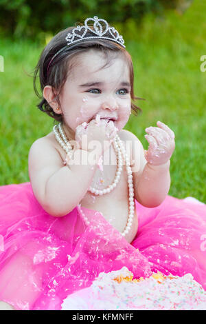 Smiling happy baby toddler girl first birthday anniversary party. Licking hand with face dirty from pink cake. Princess tiara costume Stock Photo