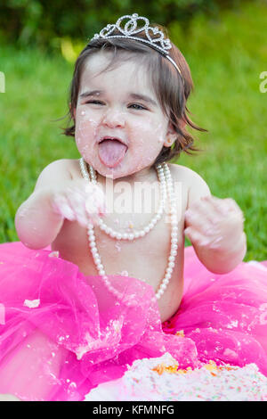 Smiling happy baby toddler girl first birthday anniversary party. Laughing and sticking tongue out, face dirty from pink cake. Princess tiara costume Stock Photo