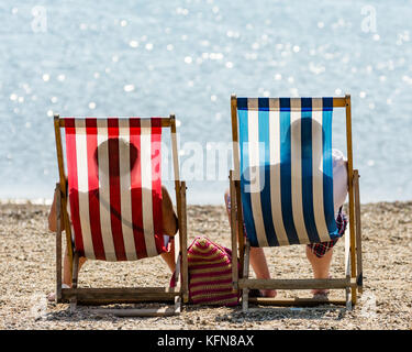 Two people sitting in deckchairs on the beach, photographed from behind, their shadows clear on the fabric - red white and blue colours Stock Photo
