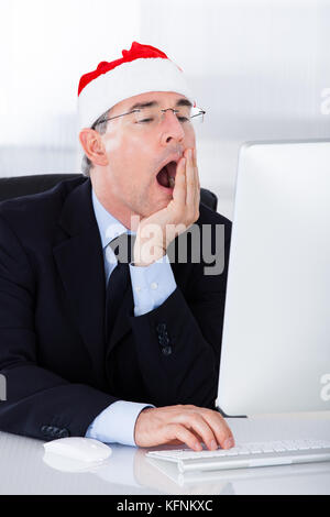 Mature Businessman With Santa Hat Yawning While Working On Computer Stock Photo