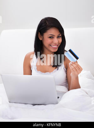 Young Woman Sitting On Bed And Shopping Online Stock Photo