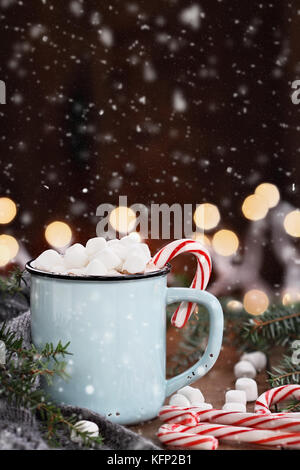 Enamel cup of hot cocoa with mini marshmallows and candy canes with pine boughs and gray scarf against a rustic background with beautiful Christmas li Stock Photo