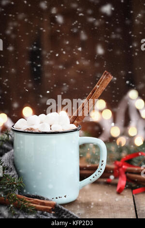 Enamel cup of hot cocoa with mini marshmallows and cinnamon bark.  Pine boughs and gray scarf against a rustic background with beautiful Christmas lig Stock Photo