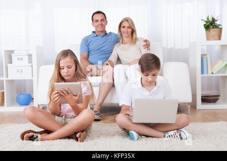 Children On The Carpet Using Tablet And Laptop With Parents Behind Them At Home Stock Photo