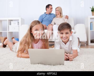 Children On The Carpet Using Tablet And Laptop With Parents Behind Them At Home Stock Photo