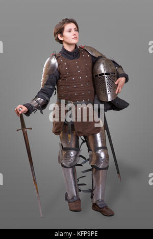 young woman in Knight Armour holding helmet in one hand and sword on her shoulder isolated over grey background Stock Photo