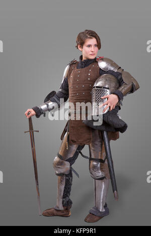 young woman in Knight Armour holding helmet in one hand and sword on her shoulder isolated over grey background Stock Photo