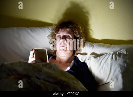 Middle aged woman reading her Apple iPhone smartphone in bed at night Stock Photo