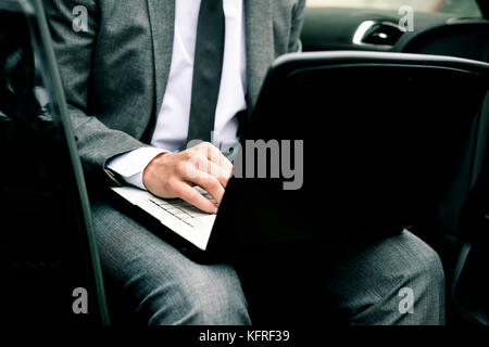 closeup of a young caucasian businessman in a gray suit using a laptop sitting at the back seat of a car Stock Photo