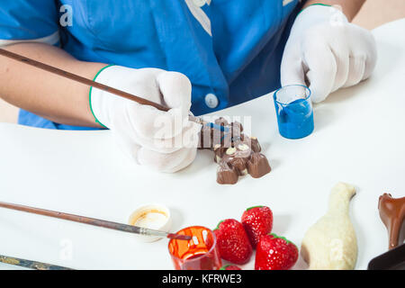 A woman confectioner with blue uniform and white sterile gloves colors a dog-shaped candy in golden color with edible paint on a white table for a New Stock Photo