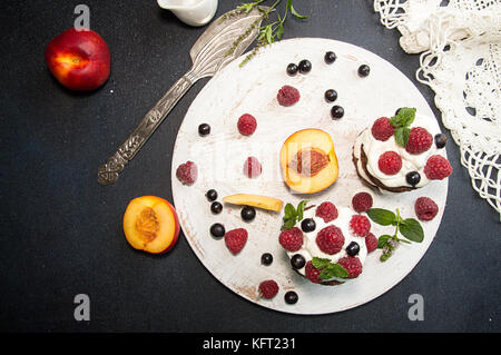 Chocolate cake decorated with raspberry, black currant, nectarine, wipped cream and mint on white wooden board on dark background. Top view. Copy spac Stock Photo
