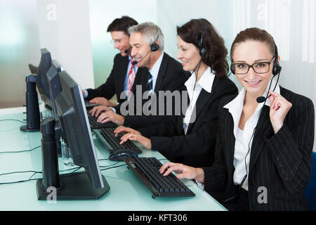 Line of professional stylish call centre operators wearing headsets seated behind their computers giving assistance Stock Photo