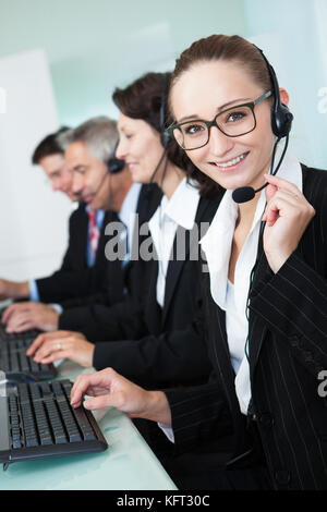Line of professional stylish call centre operators wearing headsets seated behind their computers giving assistance Stock Photo