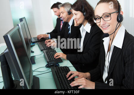 Line of professional stylish call centre operators wearing headsets seated behind their computers giving assistance Stock Photo