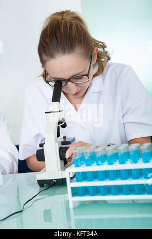 Female laboratory technician using a monocular microscope to read test results which she is recording on paper Stock Photo