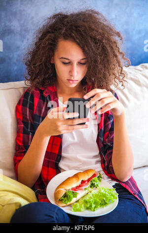 Curly hair teen girl using mibile phone and eating sandwich on sofa Stock Photo