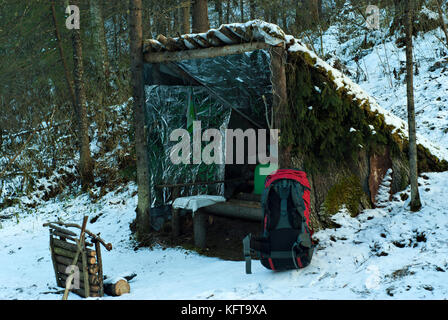 Improvised, deliberately primitive lean-to shelter from poles, bark and branches in the winter snow-covered forest. In the foreground is a red modern  Stock Photo
