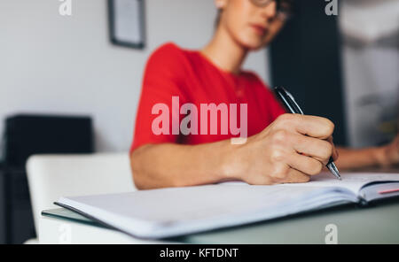 Woman wearing spectacles sitting at her desk in office and making notes in a diary. Closeup of a business woman writing in diary. Stock Photo