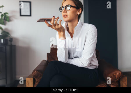 business woman talking over mobile phone on loudspeaker sitting on a couch. Woman in formal business attire holding a mobile phone close to her mouth Stock Photo