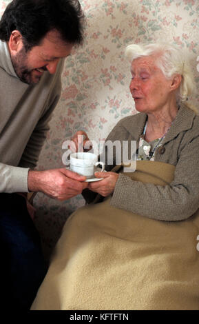 man/carer/son handing cup of tea to geriatric lady in nursing home Stock Photo