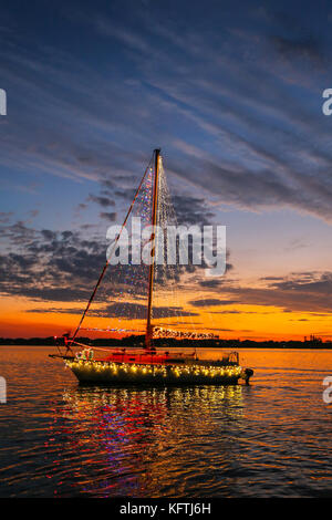 A sailboat decorated with Christmas lights passes in front of bright sunset Stock Photo