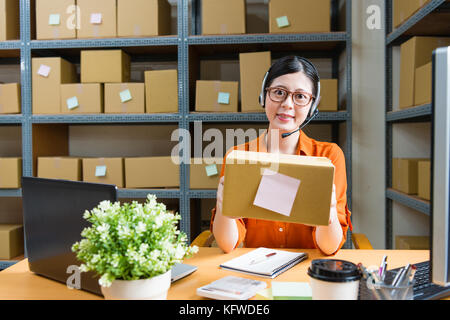 confident smiling office worker woman holding online shopping order parcel and wearing earphone sitting in warehouse office looking at camera. Stock Photo