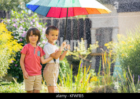 Two adorable children, boy brothers, playing with colorful umbrella under sprinkling water in their backyard Stock Photo
