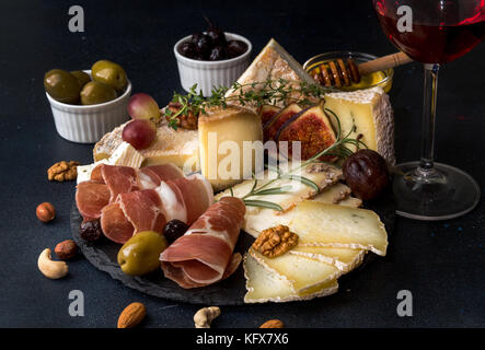 Glass of red wine and cheese plate with pieces moldy cheese, prosciutto, pickled plums, olives, grapes, figs, honey, pear, nuts on black slate backgro Stock Photo