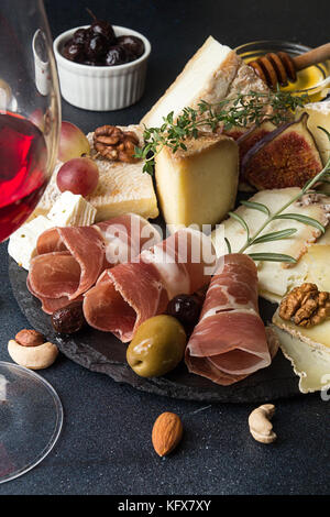 Glass of red wine and cheese plate with pieces moldy cheese, prosciutto, pickled plums, olives, grapes, figs, honey, pear, nuts on black slate backgro Stock Photo